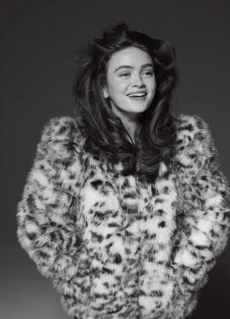 Portrait of a smiling young woman with long wavy brown hair wearing a patterned faux fur coat in a studio setting, captured in black and white.