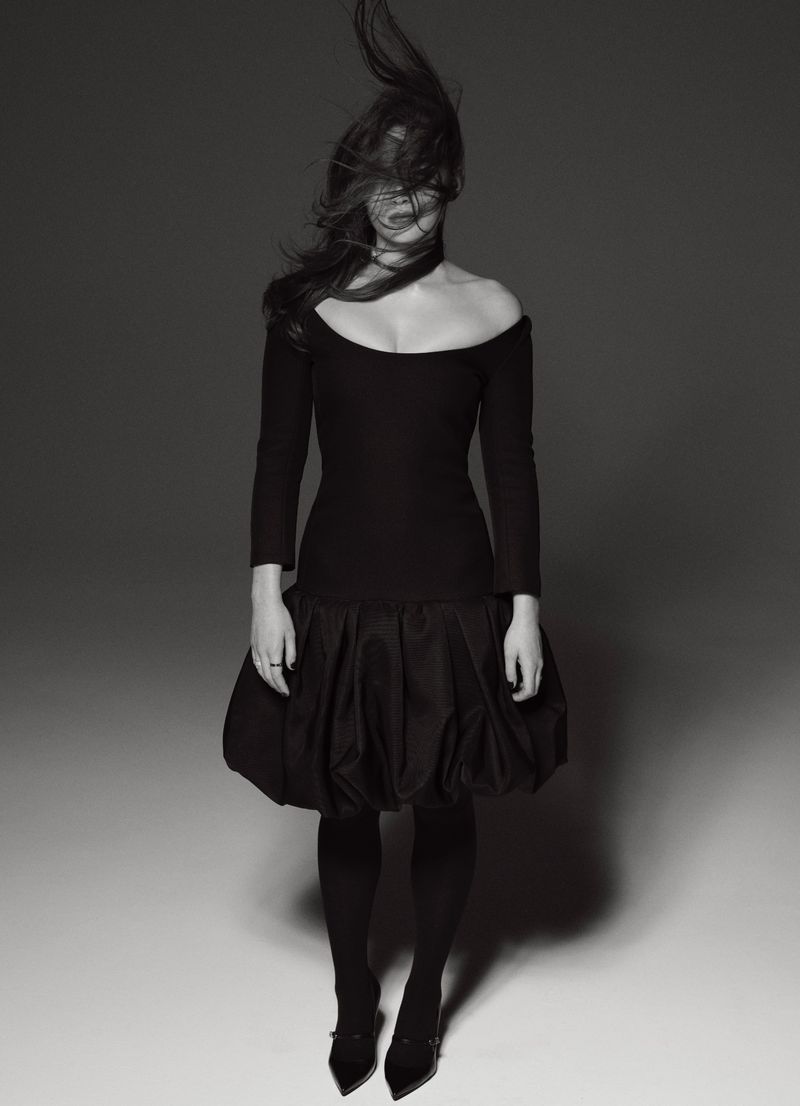 A woman in a black dress stands in a photo studio with her dark hair partially obscuring her face, creating a dramatic, artistic image.