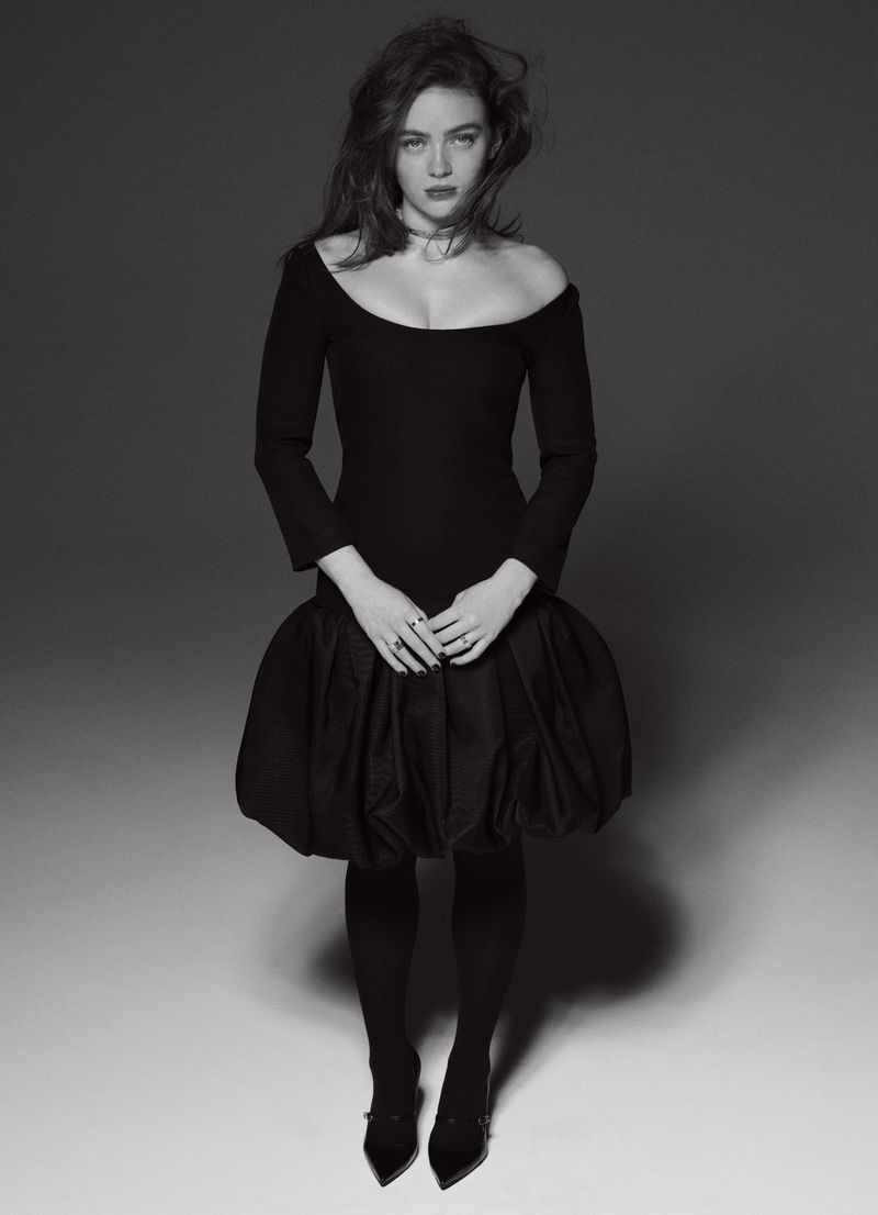 Monochrome portrait of a young woman in a black dress with a puffed skirt, black tights, and heels against a grey backdrop.