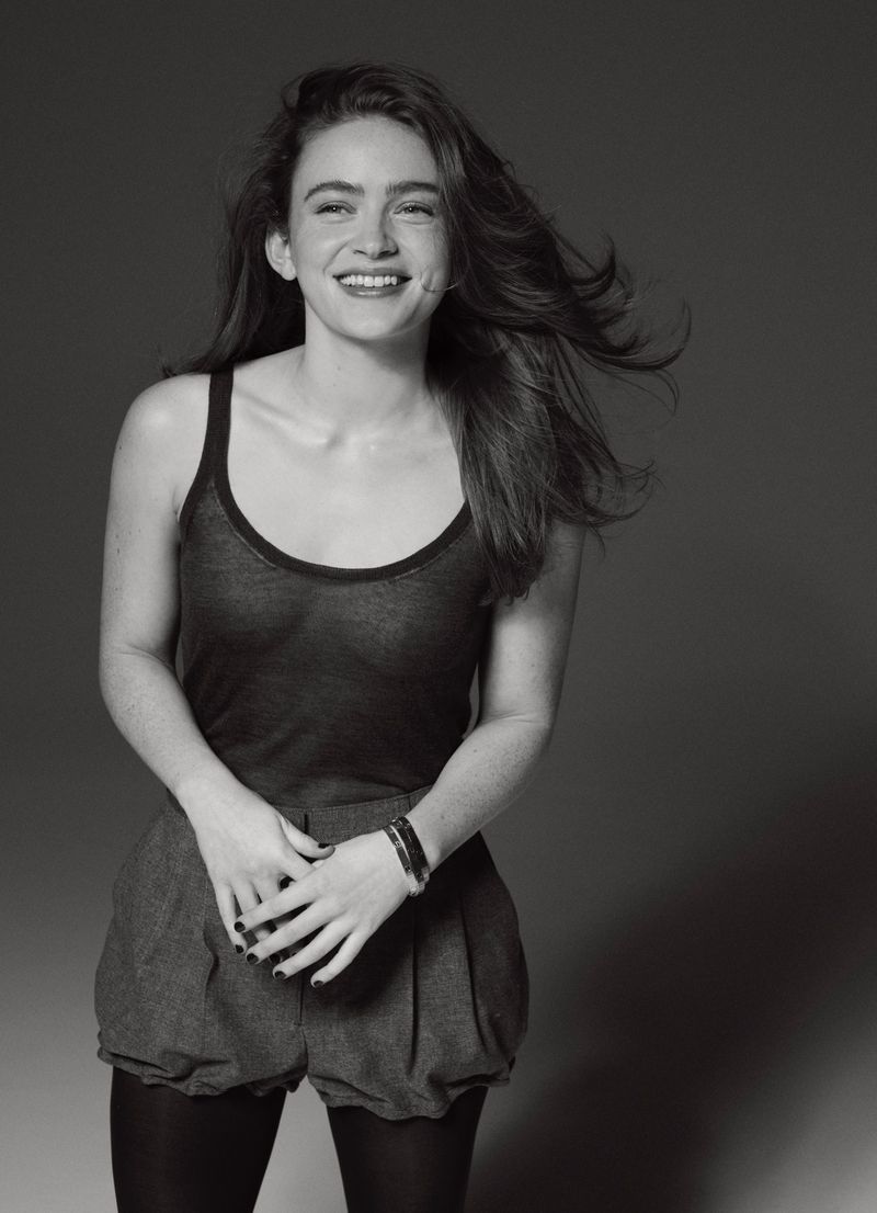 Portrait of a smiling woman with long brown hair wearing a dark tank top and shorts, standing against a neutral gray backdrop.