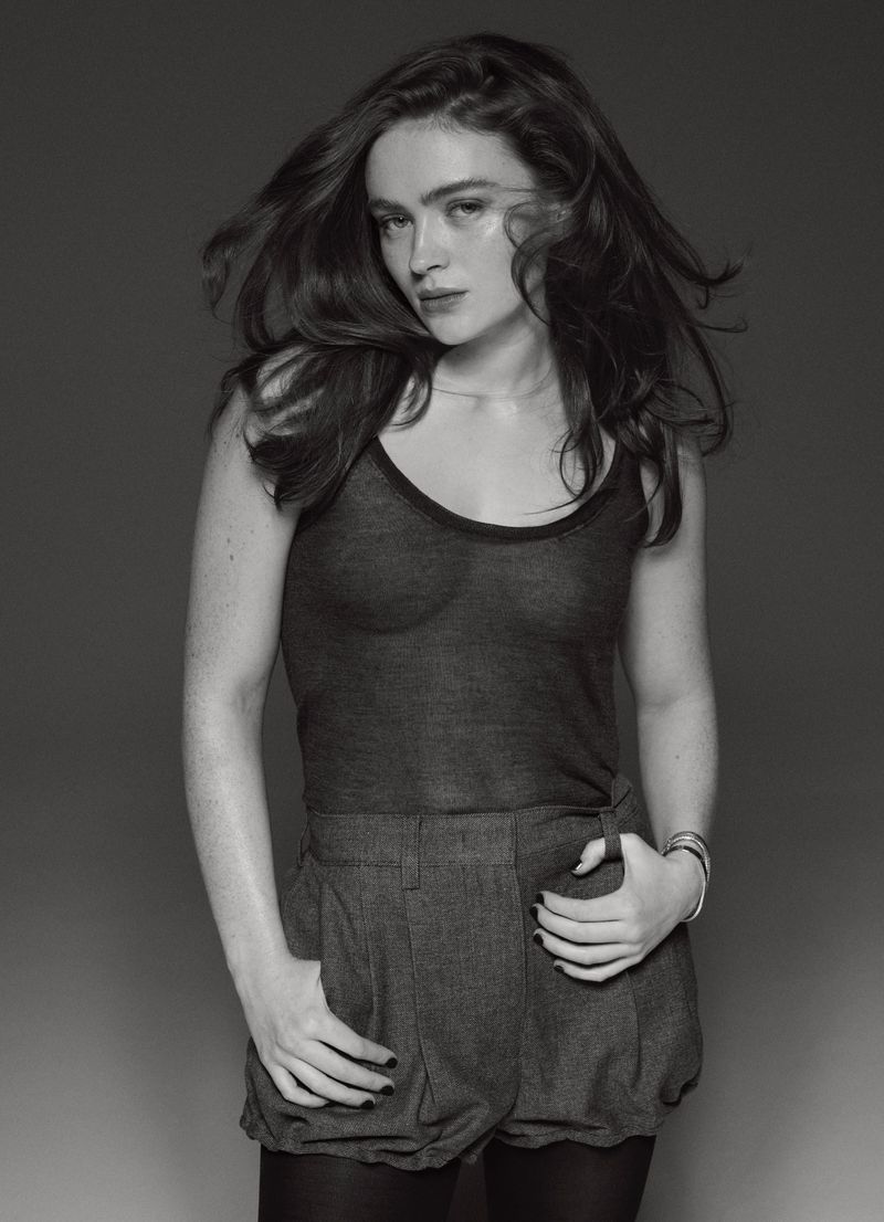 Black and white portrait of an adult female fashion model with long wavy hair wearing a tank top and shorts with grey background.