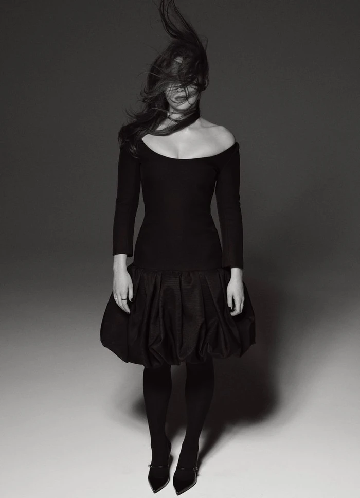 Portrait of Sadie Sink wearing a black dress with blown hair against a grey seamless studio background.