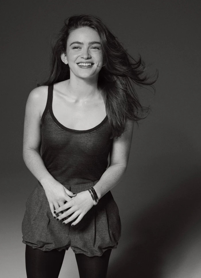 Portrait of Sadie Sink smiling in a dark tank top and shorts against a neutral studio backdrop