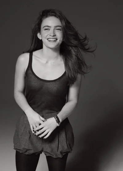 Portrait of Sadie Sink smiling in a dark tank top and shorts against a neutral studio backdrop