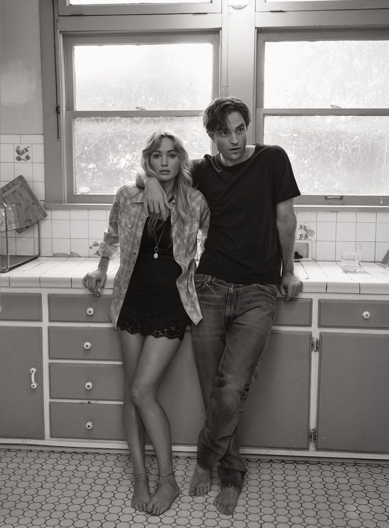 Black-and-white portrait of a stylish barefoot couple in a kitchen with natural light. The woman wears a short dress and unbuttoned shirt.
