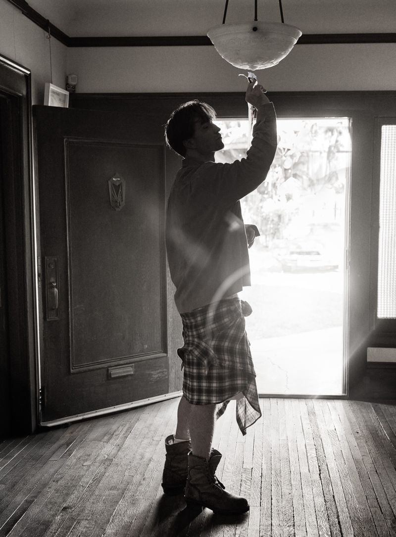 Monochrome full shot of a person indoors, reaching up to a light fixture while standing in a doorway with outdoor light beyond.