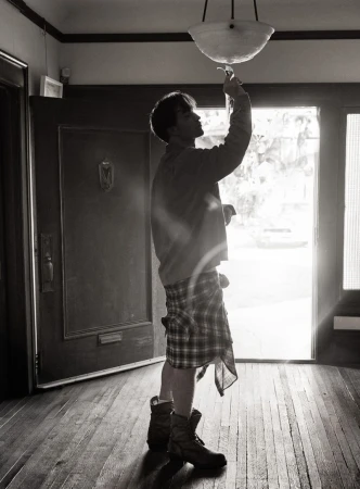 Monochrome full shot of a person indoors, reaching up to a light fixture while standing in a doorway with outdoor light beyond.