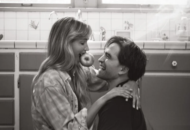 Black and white photograph of a woman with long hair biting an apple while a man behind her smiles in an indoor kitchen setting.