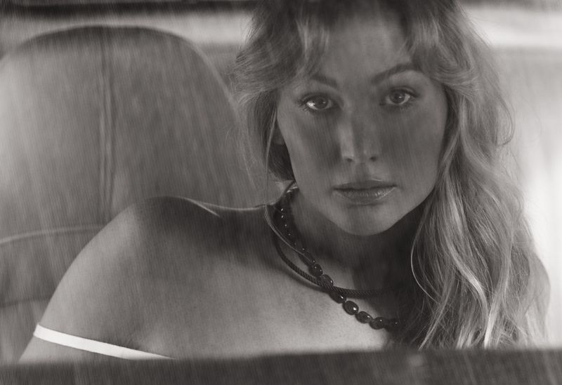Jennifer Lawrence photographed in black and white seated inside a car wearing a dark beaded necklace