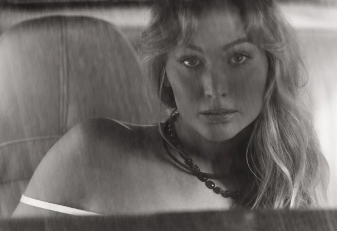 Jennifer Lawrence photographed in black and white seated inside a car wearing a dark beaded necklace