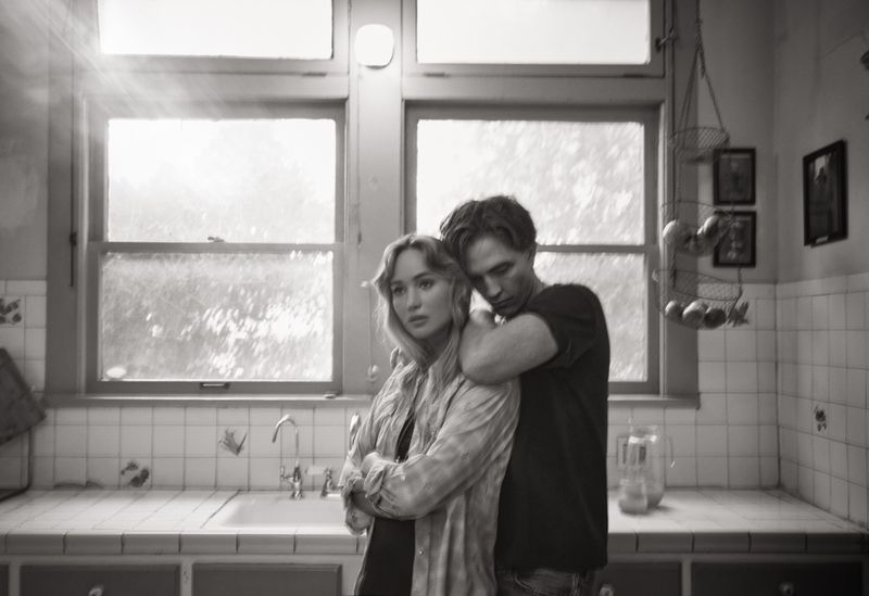 Black-and-white photo of a couple indoors: a woman in a patterned shirt standing with her arms crossed, embraced by a man in a t-shirt.