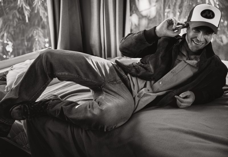 Robert Pattinson lying on a bed wearing a trucker hat and jacket for V Magazine photographed by Cass Bird