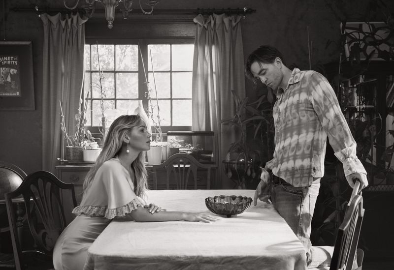 Black and white portrait of a stylish couple indoors, the woman seated at a table, the man standing, creating an intimate, vintage feel.