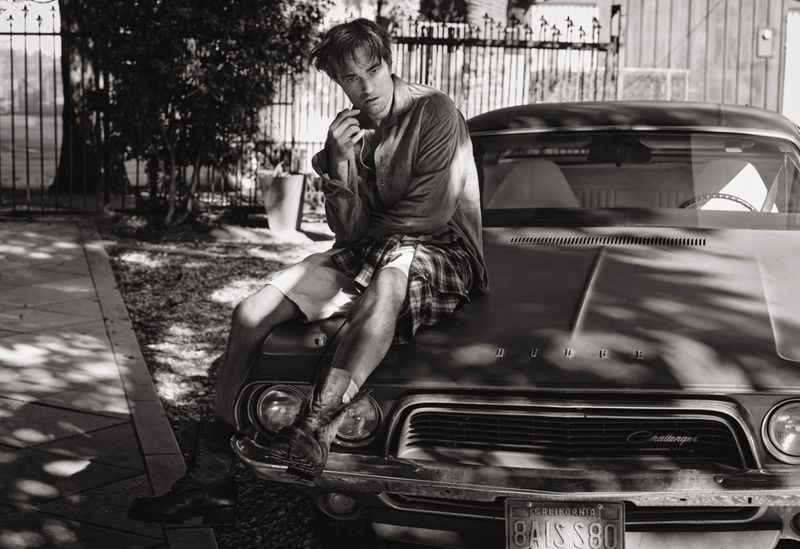 A monochrome outdoor shot of a young adult male sitting on the hood of a vintage car with a gate and trees in the background.