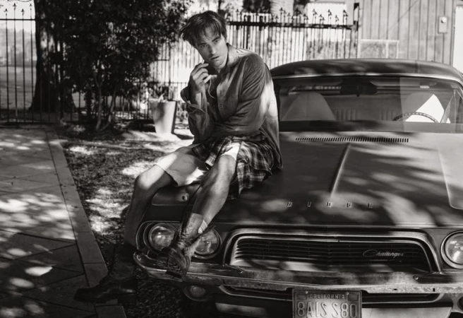 A monochrome outdoor shot of a young adult male sitting on the hood of a vintage car with a gate and trees in the background.