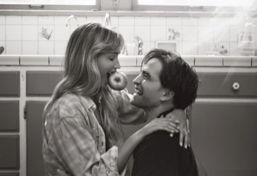 Black and white portrait of Jennifer Lawrence holding an apple between her teeth while looking at Robert Pattinson in a kitchen
