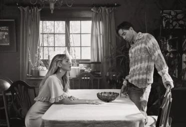 Black and white editorial portrait of Jennifer Lawrence seated at a dining table looking up at Robert Pattinson standing