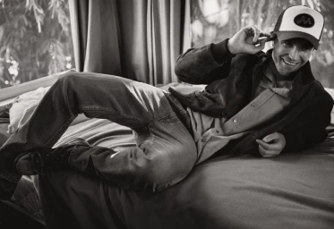 Robert Pattinson lying on a bed wearing a trucker hat and jacket for V Magazine photographed by Cass Bird