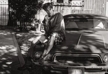 Robert Pattinson seated on the hood of a vintage car wearing shorts and a button-down shirt in a black and white editorial portrait.