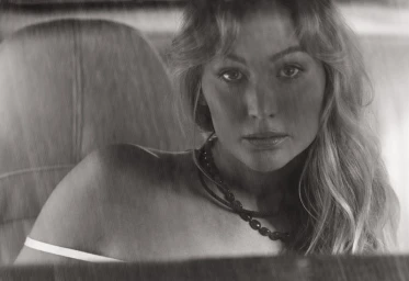 Jennifer Lawrence photographed in black and white seated inside a car wearing a dark beaded necklace