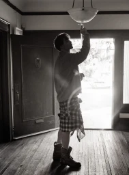 Robert Pattinson standing indoors reaching up toward a ceiling light fixture in a black and white photograph by Cass Bird for V Magazine.