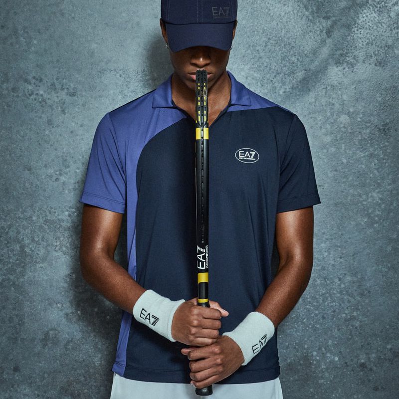 Man in athletic wear holding a tennis racket against textured gray background. Polo shirt and cap feature EA7 logo. Wristbands are also visible.