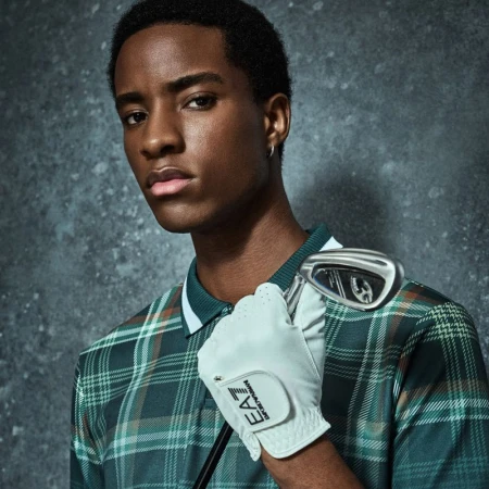 Young man in golf attire holding club gazes intensely against textured gray backdrop; fashion and sport blend in portrait.