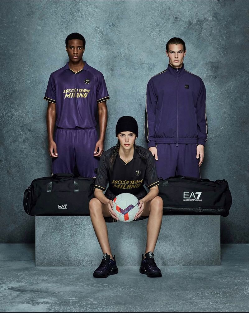 Three people showcasing soccer apparel, two standing and one sitting holding a soccer ball, with duffel bags on a grey background.