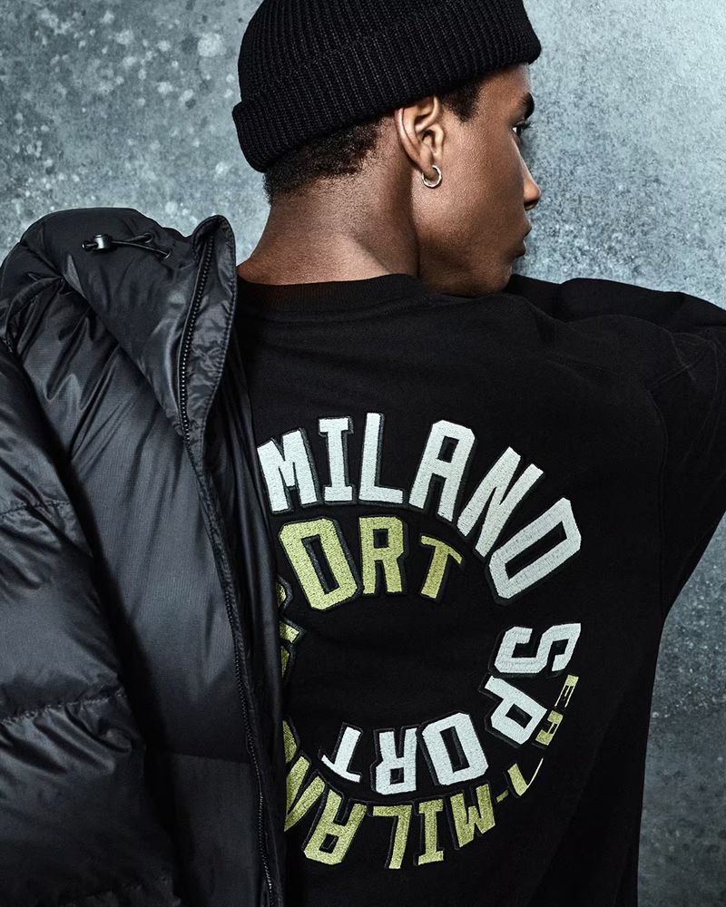A man wearing a black beanie and a black long-sleeved shirt with white lettering over a textured gray background with a black puffer coat.