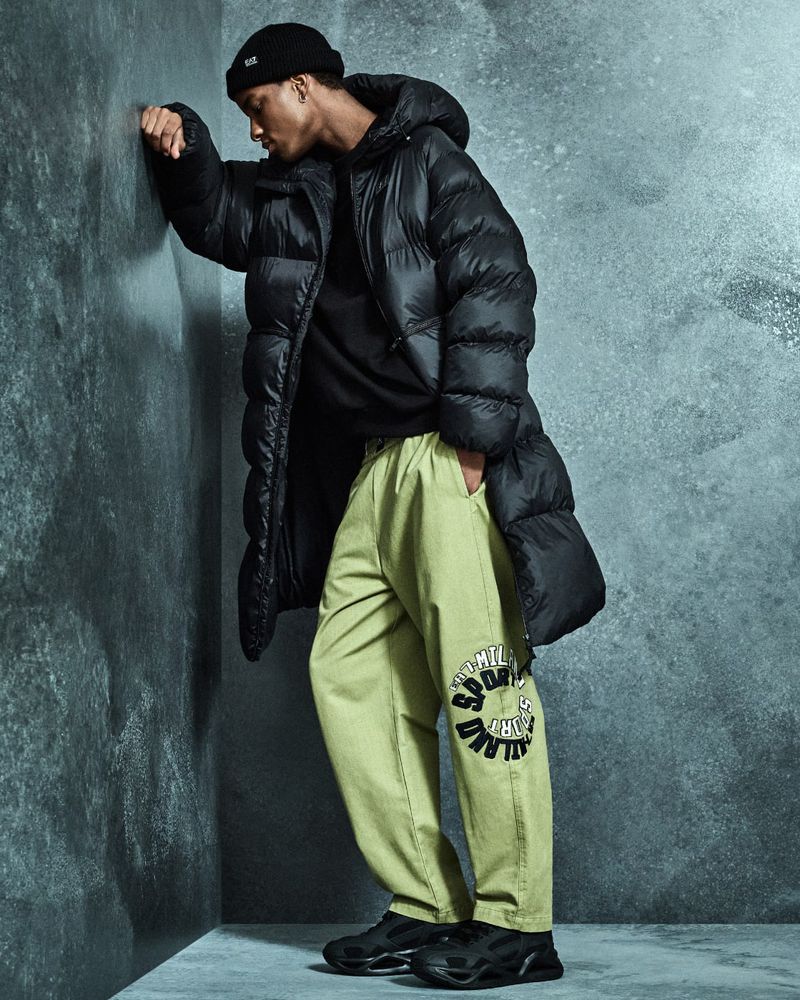 Man in a black beanie, puffer coat, and light green pants leans against a gray textured wall in a studio setting.