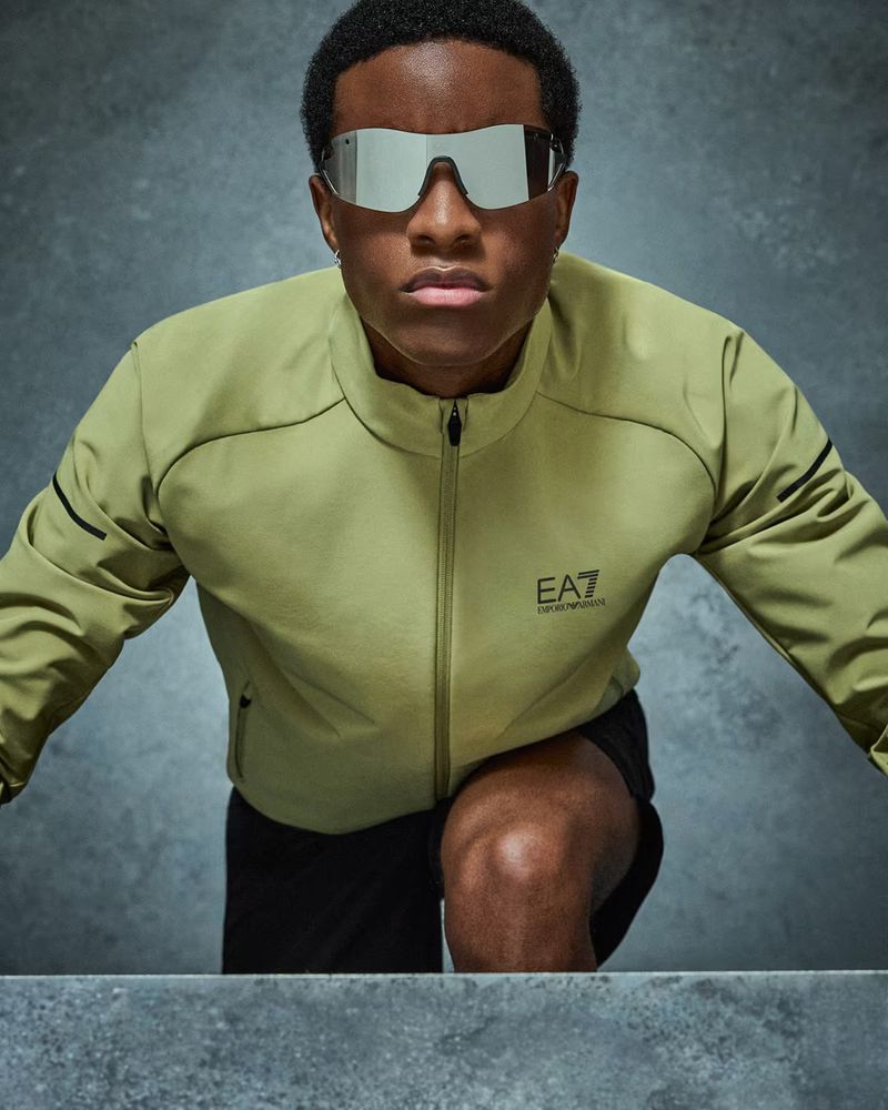 Young man poses indoors wearing stylish green jacket and mirrored sunglasses in a fashion studio setting with a neutral background.