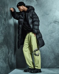 Model leaning against a textured gray wall wearing a black puffer coat and light green pants.