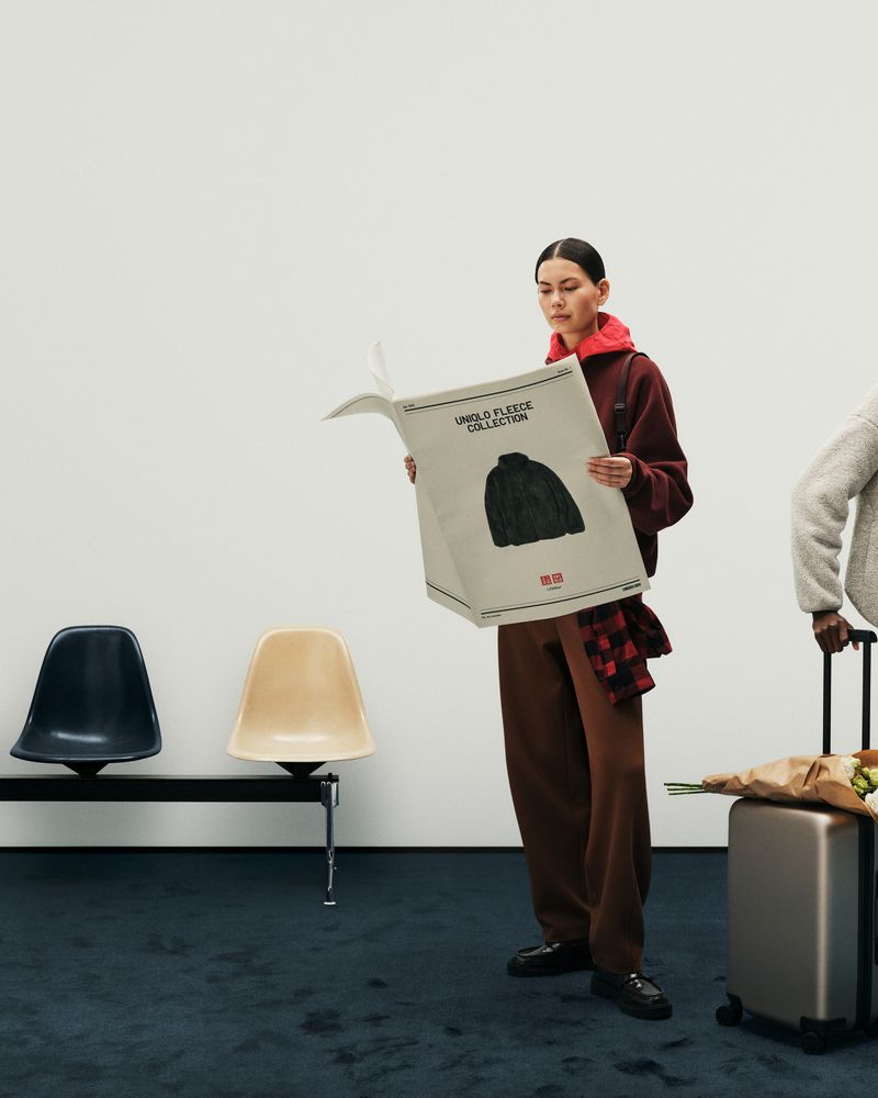 A person holds a large paper bag with a clothing graphic in an indoor setting with chairs and luggage for a fashion editorial.