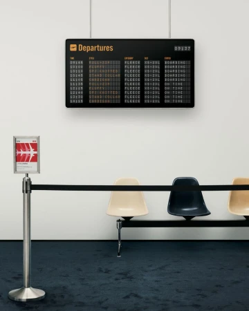 Indoor shot of an airport departures board displaying flight information, passenger queue barrier, and empty chairs against a white wall.