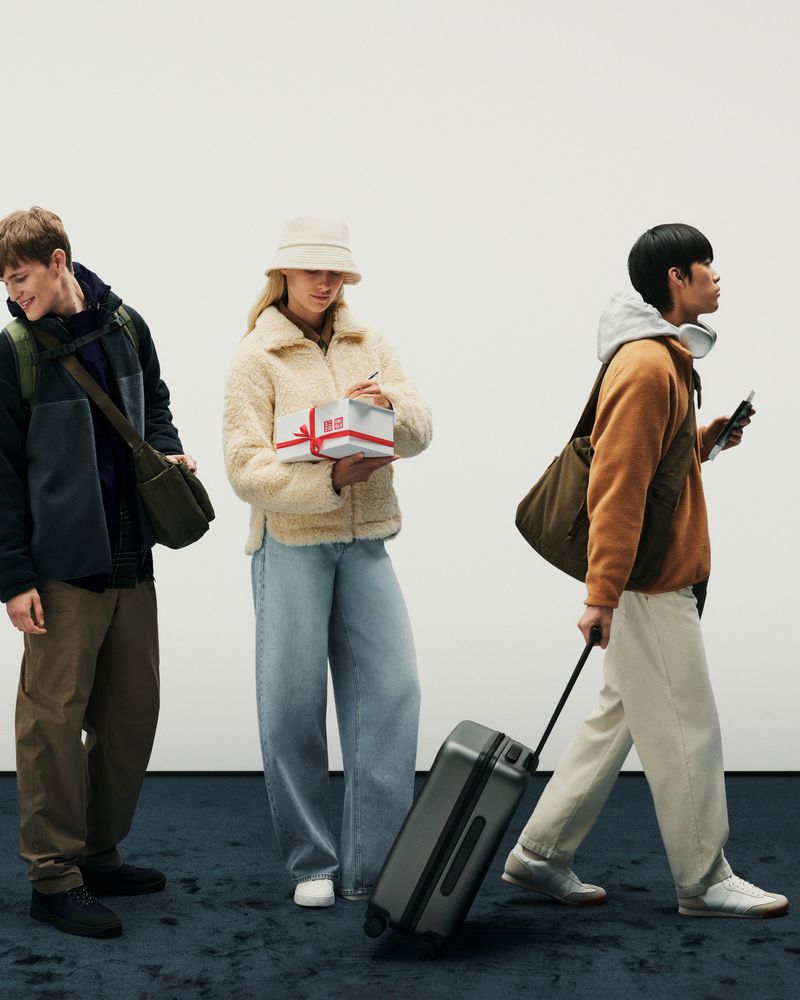 Three people standing in a row against a white wall; one rolling a suitcase, one holding a gift, and one holding a bag.