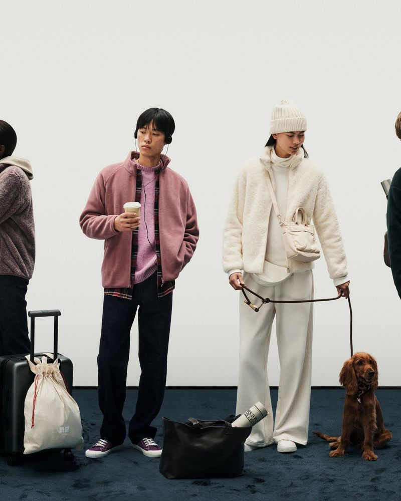Several adults standing in a row, wearing casual clothing in a studio, one woman holding a dog on a leash, and luggage at their feet.