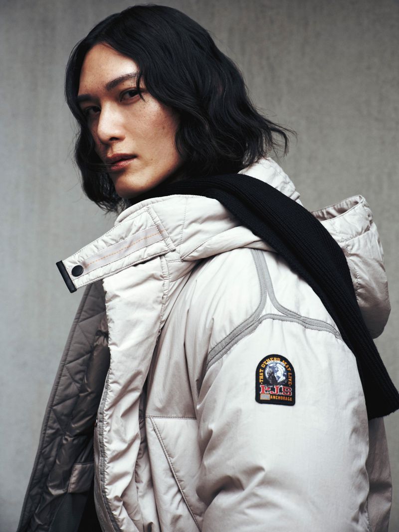 Adult male wearing a cream-colored hooded parka jacket with a black scarf against a neutral background in a studio setting.