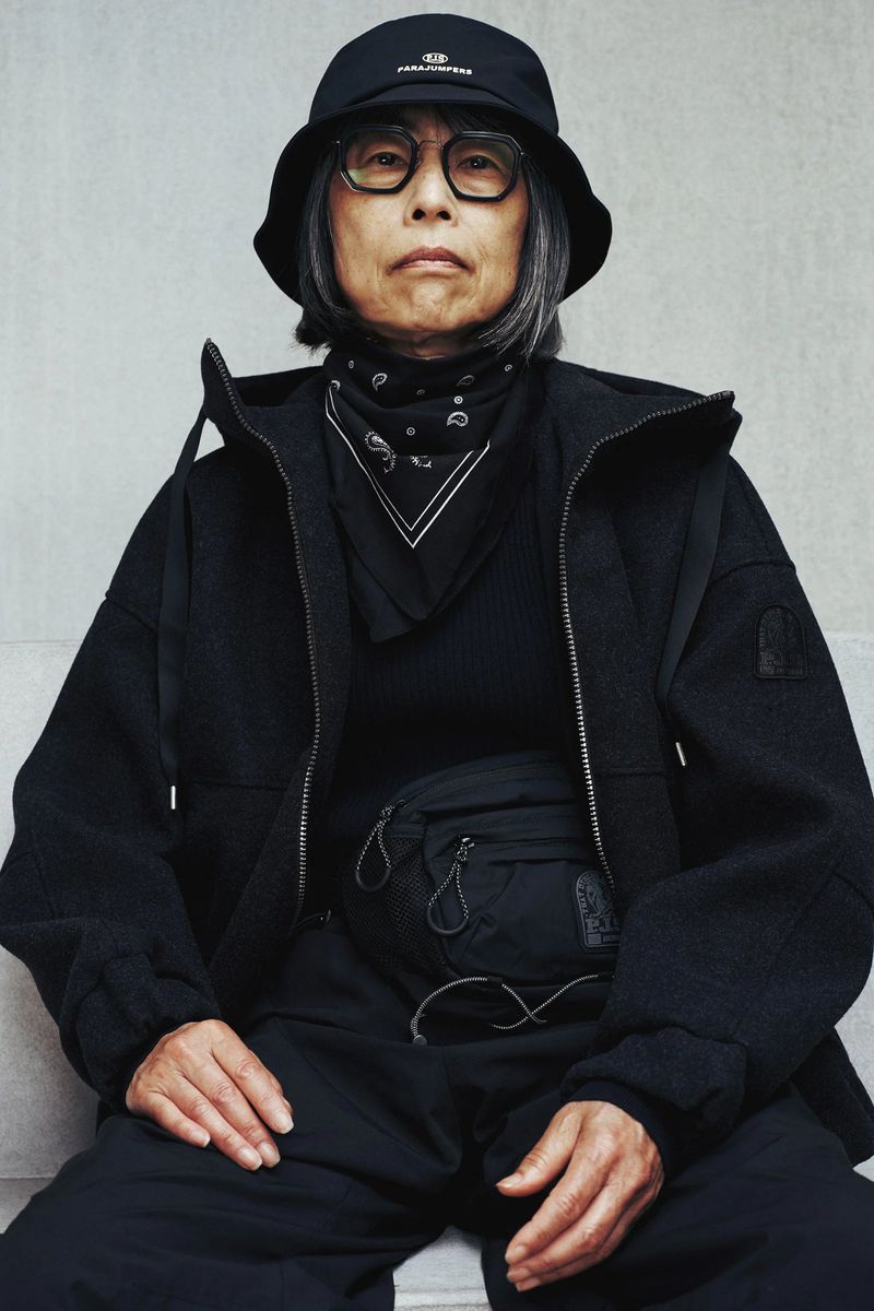 Elderly woman wearing black bucket hat and jacket poses in a studio with neutral wall background. Black fanny pack detail.