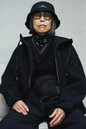 Elderly woman wearing black bucket hat and jacket poses in a studio with neutral wall background. Black fanny pack detail.