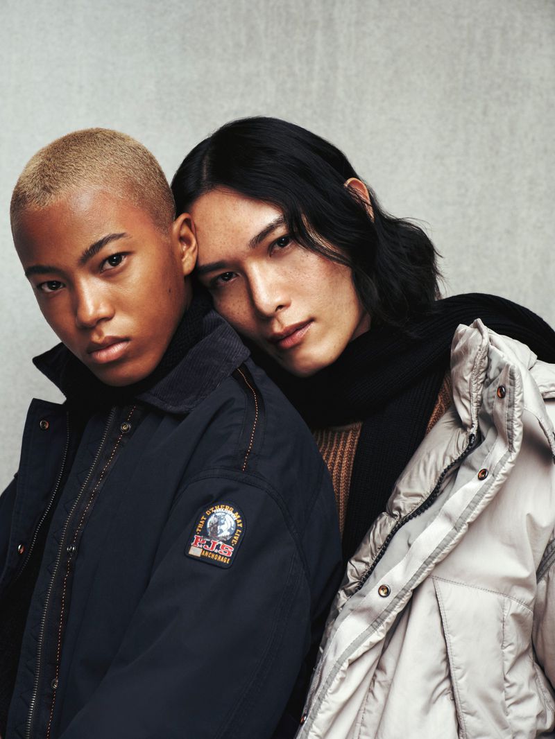 Two fashionable people pose in outerwear. Person on left wears a blue parka with logo, while the other wears a cream-colored down coat.