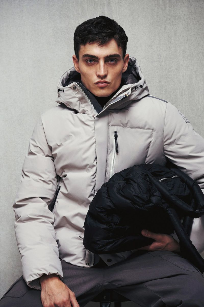 A brunette adult male model in a grey puffer coat sits in a studio with a black folded coat in his lap. Fashion shot.