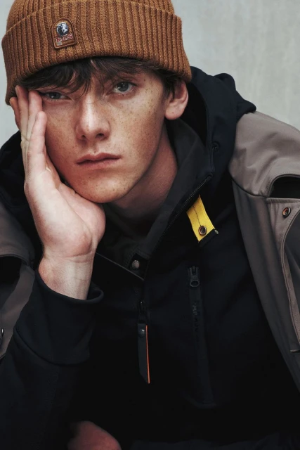 Portrait of a light-skinned adult male with brown hair, wearing a brown beanie and dark jacket, looking at the camera with a serious expression.