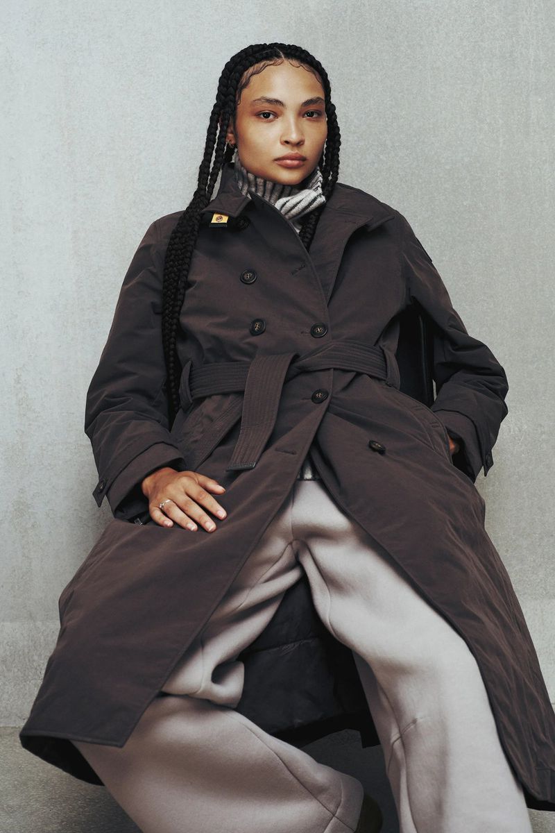Woman in brown double-breasted trench coat and grey sweatpants sitting indoors against a concrete wall in a fashion editorial.