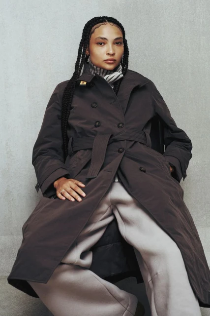 Woman in brown double-breasted trench coat and grey sweatpants sitting indoors against a concrete wall in a fashion editorial.