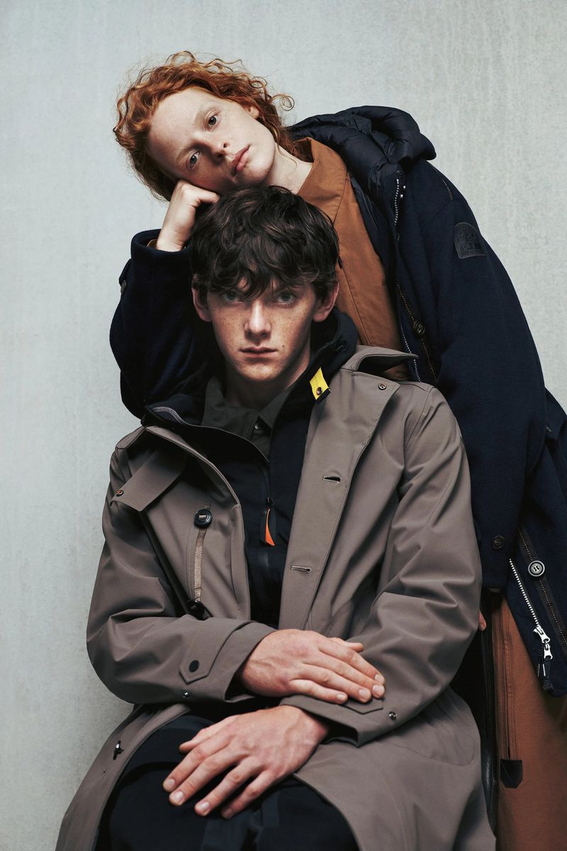 Two young adult men wearing winter coats pose indoors against a neutral wall in a fashion editorial shot in soft studio lighting.