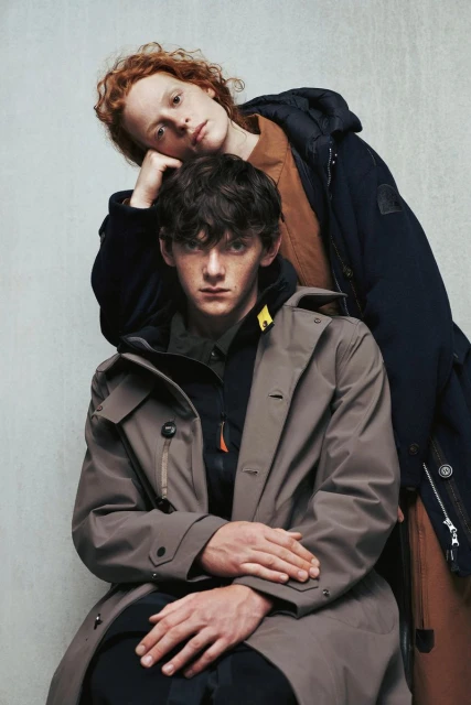 Two young adult men wearing winter coats pose indoors against a neutral wall in a fashion editorial shot in soft studio lighting.
