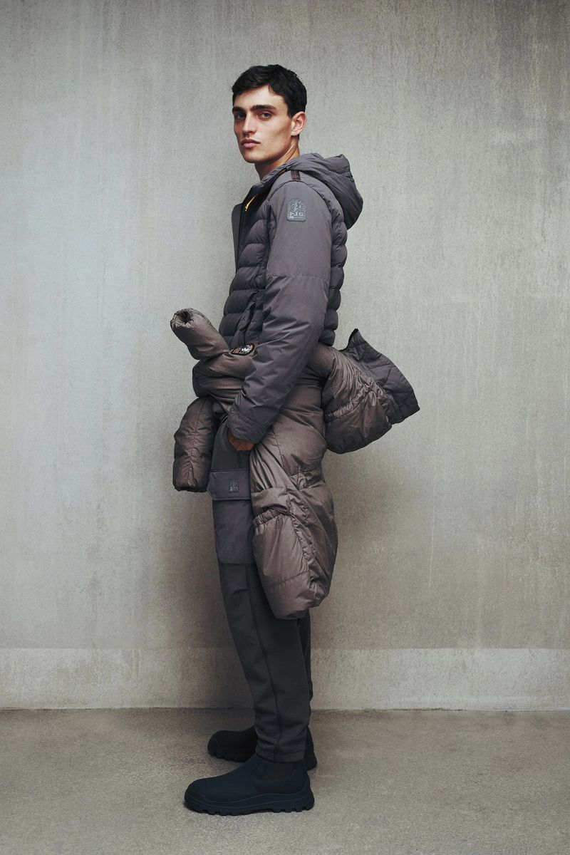 A young male fashion model poses in a grey quilted puffer jacket and pants, holding another puffer jacket in a photo studio setting.