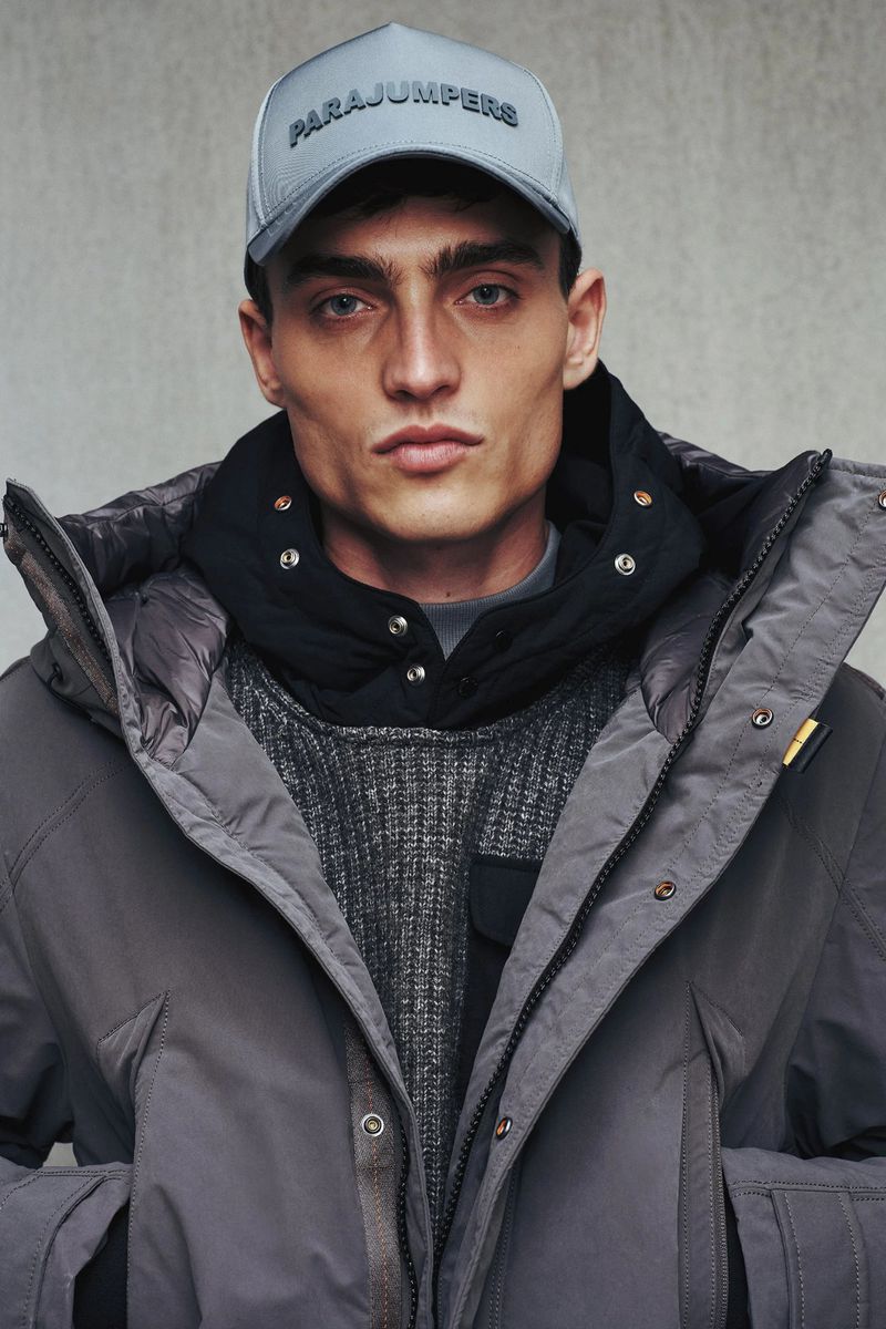 Model wearing a grey cap with branded text, a black hoodie, and a grey winter parka against a neutral concrete background