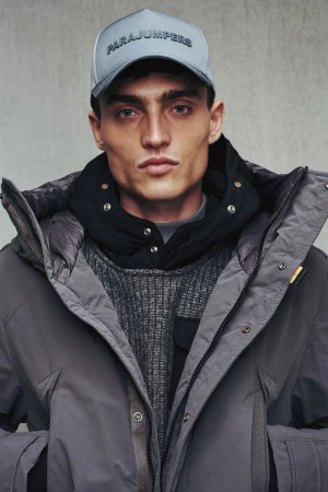 Model wearing a grey cap with branded text, a black hoodie, and a grey winter parka against a neutral concrete background