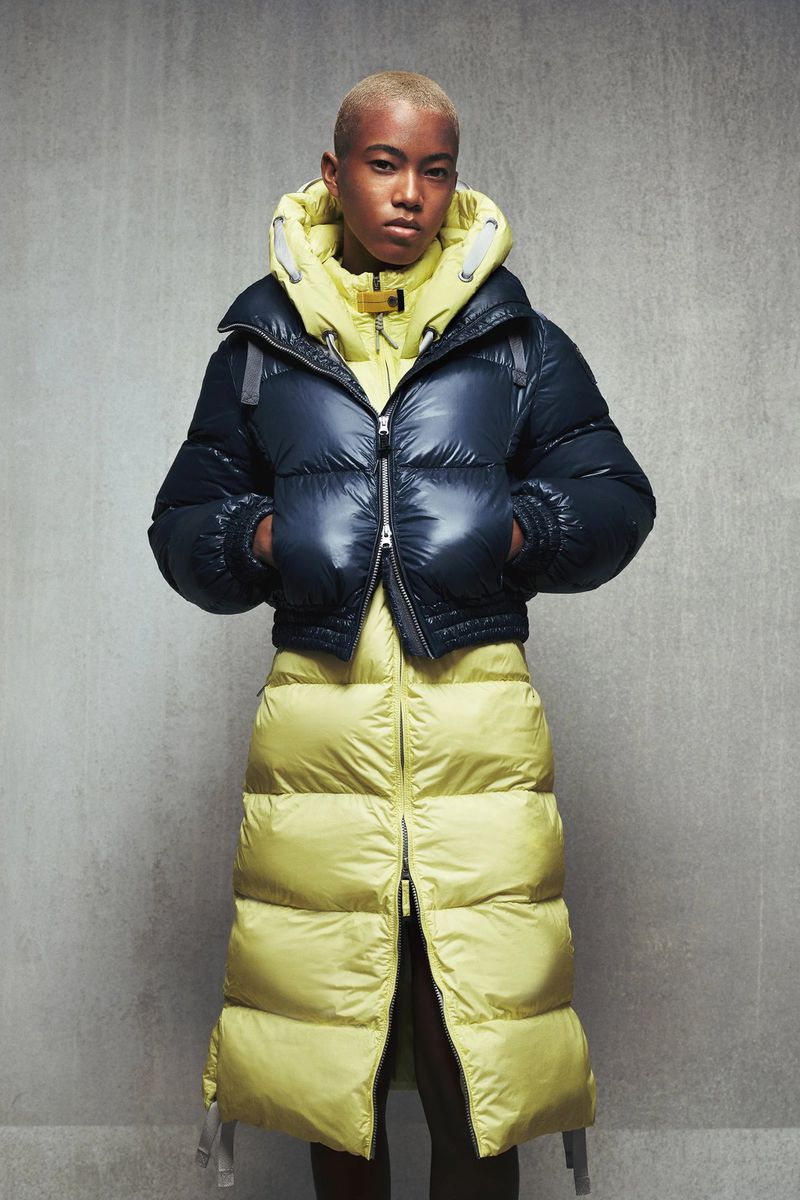 A woman with short hair poses in a two-tone navy and chartreuse puffer coat in front of a textured gray wall in a studio shot.